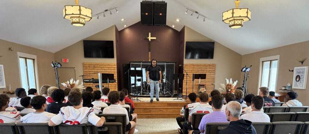 A young man stands in the front of a church and speaks to football players seated in the sanctuary.