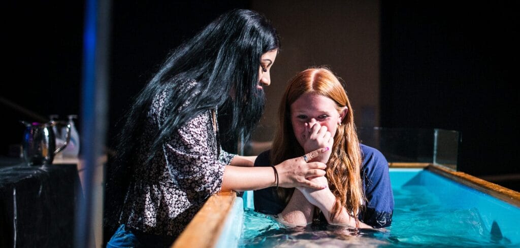 A young woman sits in a baptistry while another young woman kneels next to her, preparing to baptize her.
