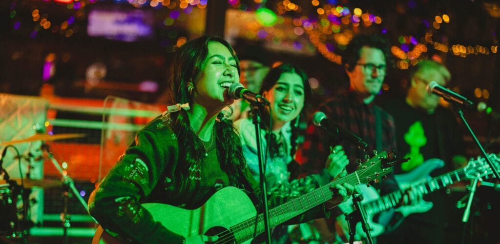 A group of musicians sing and play guitar on a stage inside a bar at Christmastime.